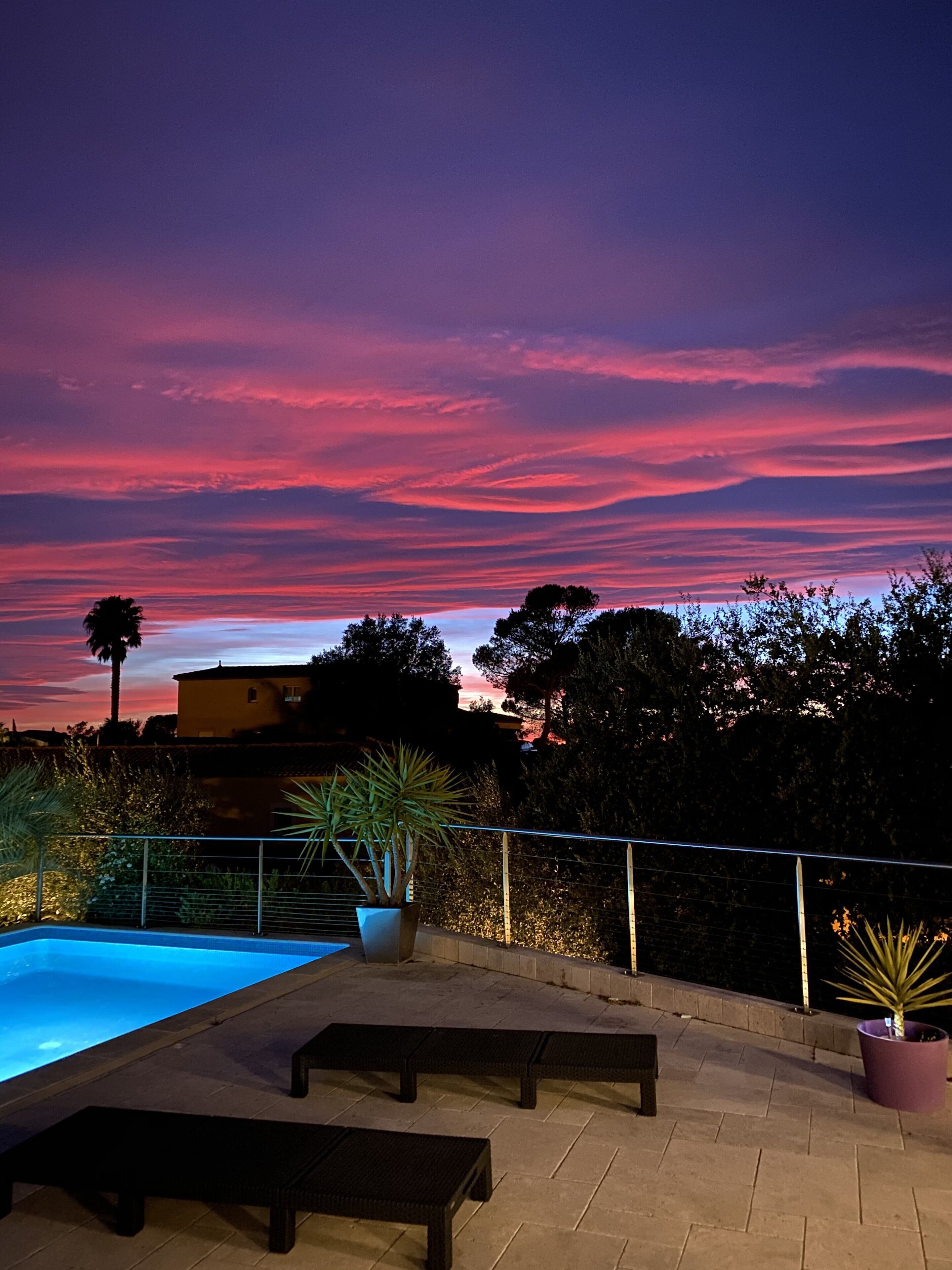 A rooftop pool area with a view of the sky at sunset, surrounded by trees and a building.
