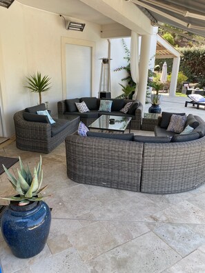 A covered outdoor seating area with wicker furniture, a glass coffee table, and potted plants.