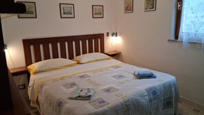 A neatly made bed with a quilt, pillows, and a folded towel. There is a bedside table with a lamp, and wall-mounted artwork above the bed. - Villa Giuliana, small two-room apartment 3/4 Ptl. (Capoliveri (LI))