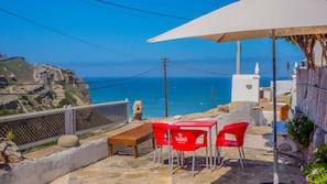 A terrace with a table and chairs, overlooking a coastal view with a clear sky.
