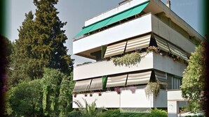 Un edificio moderno de varios pisos con balcones, rodeado de vegetación y automóviles estacionados.