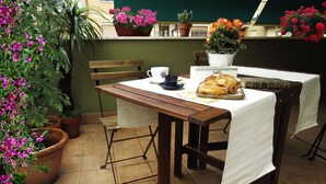 Una mesa de madera con mantel blanco, un libro y un pastel. Se ven plantas en macetas y un banco al fondo.