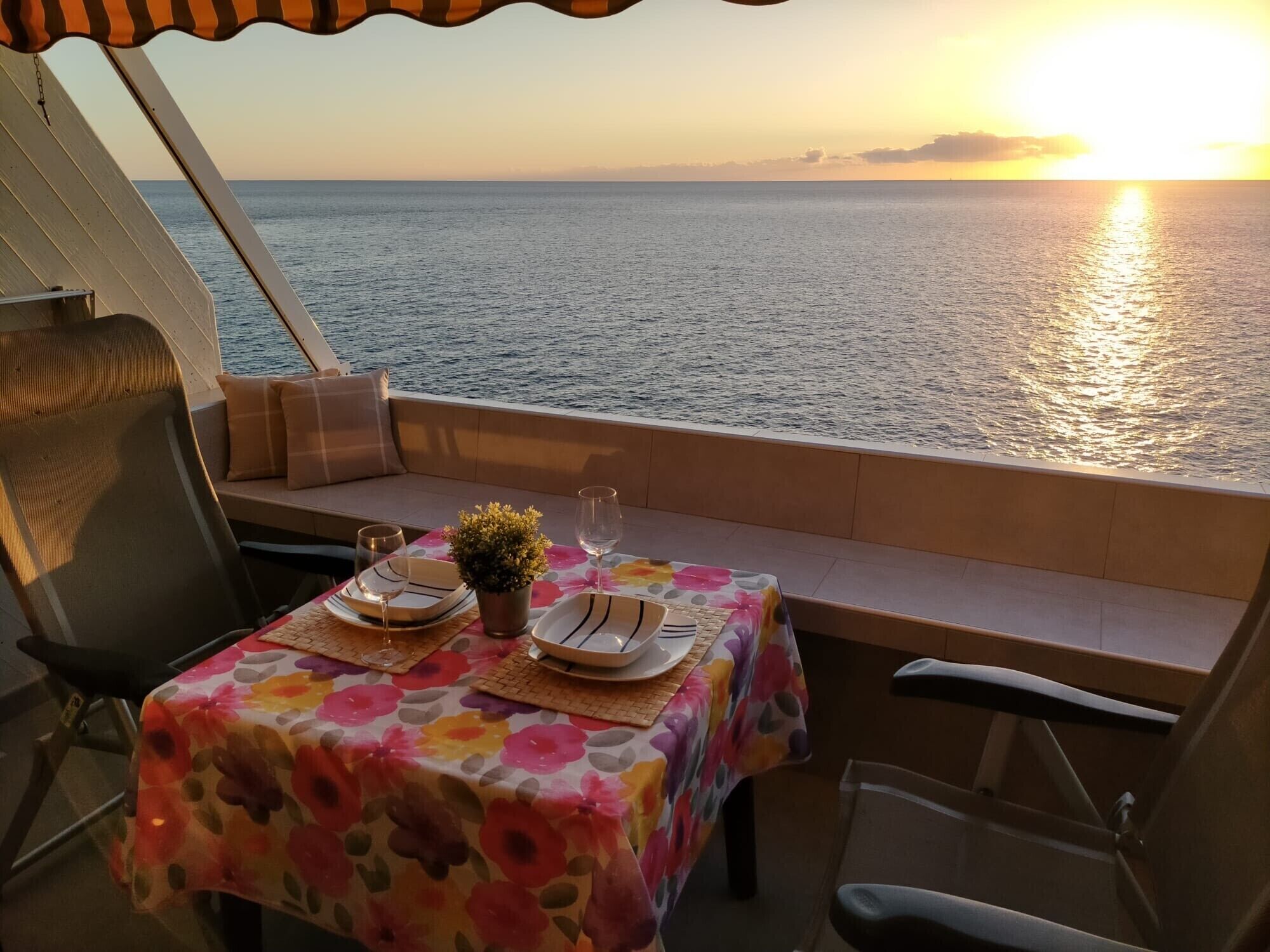 A table set for two with a floral tablecloth, a small potted plant, and a glass of water on a boat deck overlooking the sea at sunset.