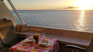 Mesa para dos con mantel floral, una pequeña planta en maceta y un vaso de agua en la cubierta de un barco, con vista al mar al atardecer.