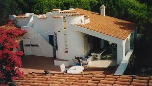Un bâtiment blanc avec un toit en tuiles, un petit patio avec une table et des chaises, et une vue sur les montagnes en arrière-plan.