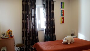 Une chambre avec deux lits, un bureau avec des jouets, une chaise, un radiateur et une fenêtre avec des rideaux.
