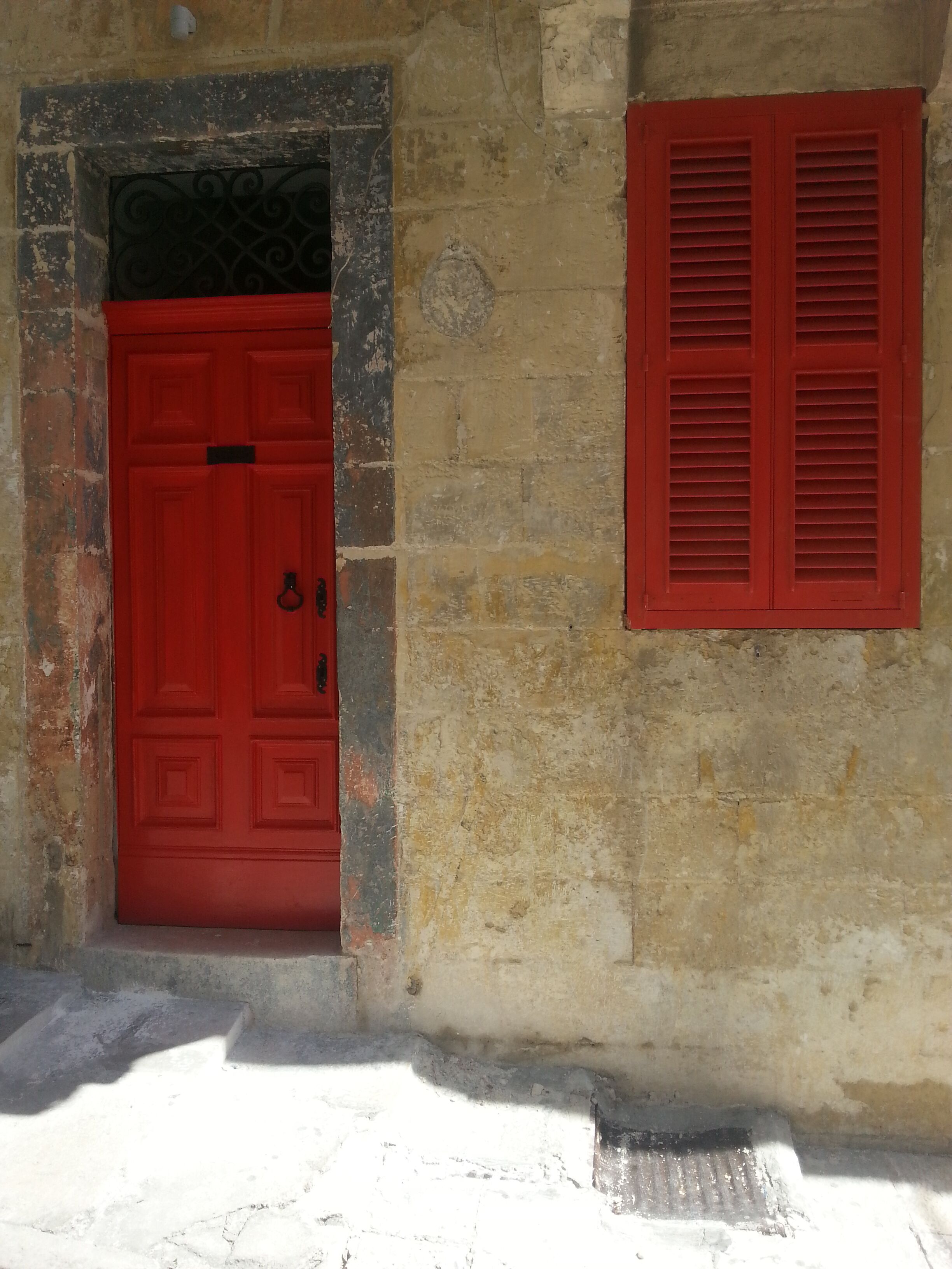A red door with a metal grille and a red shutter on a weathered stone wall.