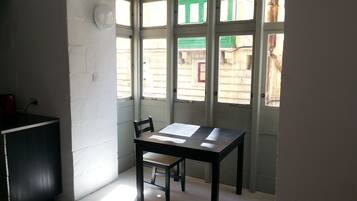 A small, sparsely furnished room with a single table and chair, large windows with green frames, and a white wall.