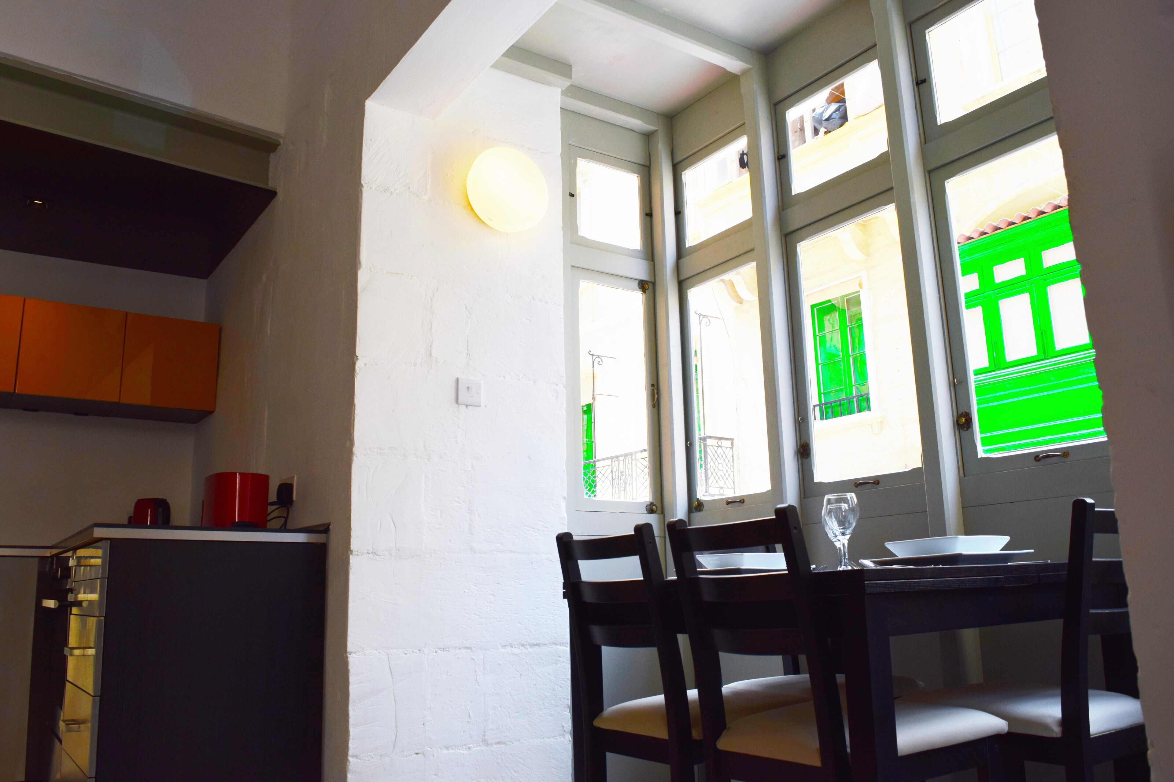 Un coin repas avec une table et des chaises, une fenêtre avec des volets verts, et un espace cuisine avec des placards et un réfrigérateur.