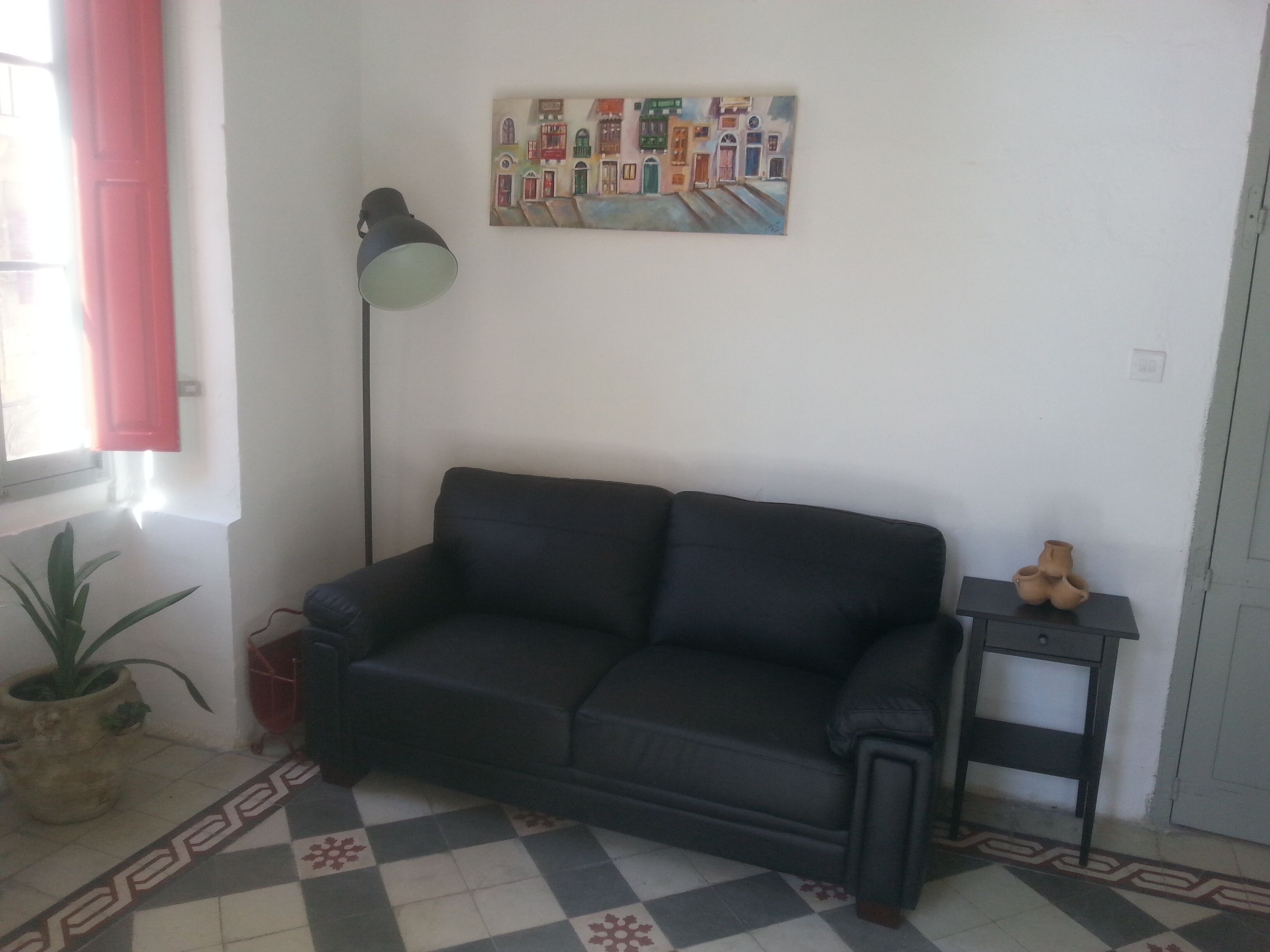 A room with a black sofa, a floor lamp, a potted plant, a small table with a vase, and a painting on the wall.
