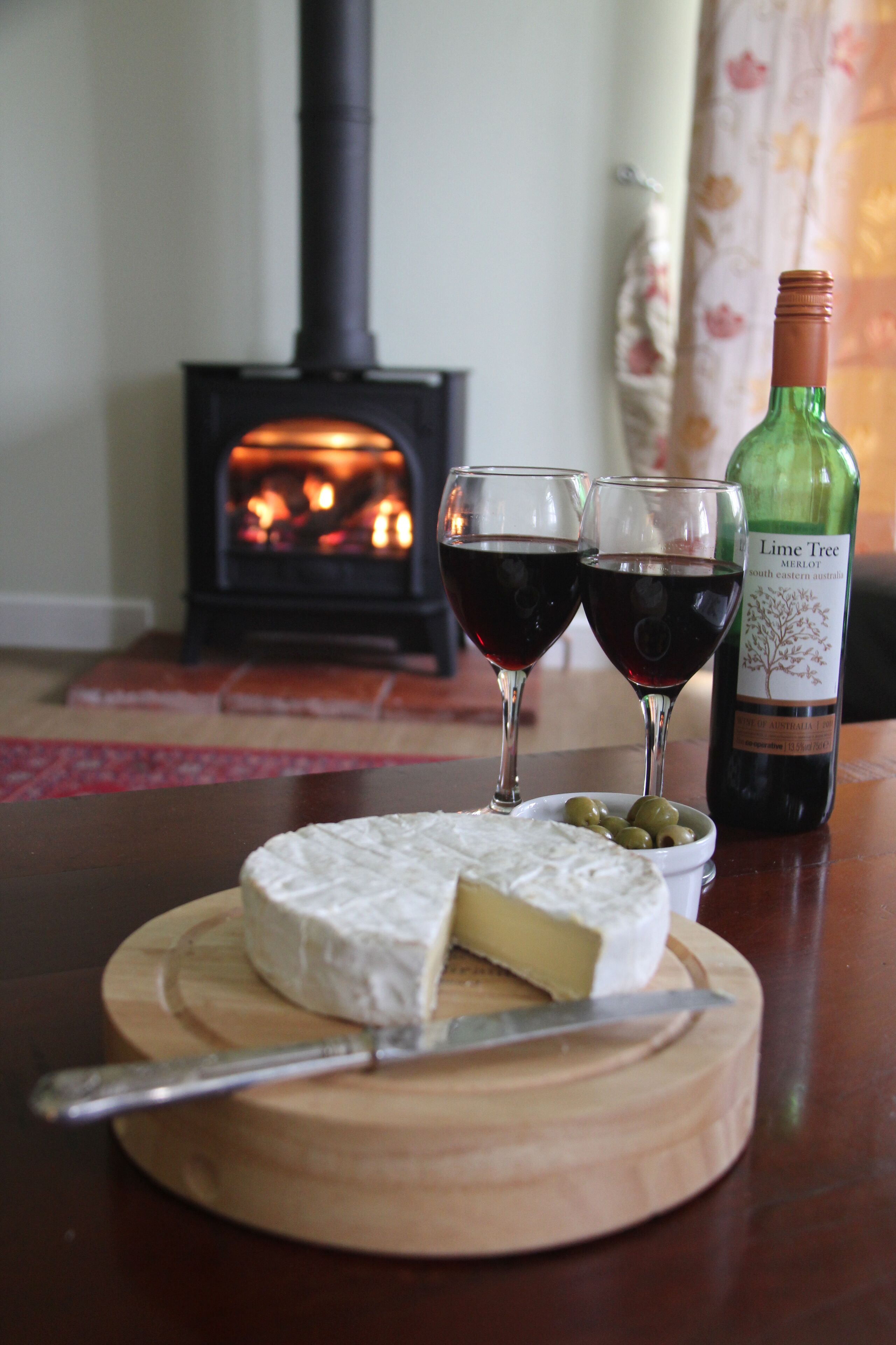 Une table en bois sur laquelle se trouvent une grande tranche de fromage, deux verres de vin rouge et une bouteille de vin.