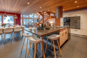 A modern kitchen with a central island, bar stools, a staircase, and a view of the outdoors. - Serre-Chevalier: 5-star luxury chalet, Spa area, panoramic view (La Salle-Les-Alpes)