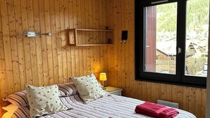 Een slaapkamer met een houten wandpaneel, een bed met gestreept beddengoed, twee kussens, een nachtkastje met een lamp en een raam met uitzicht op een gebouw en bomen.