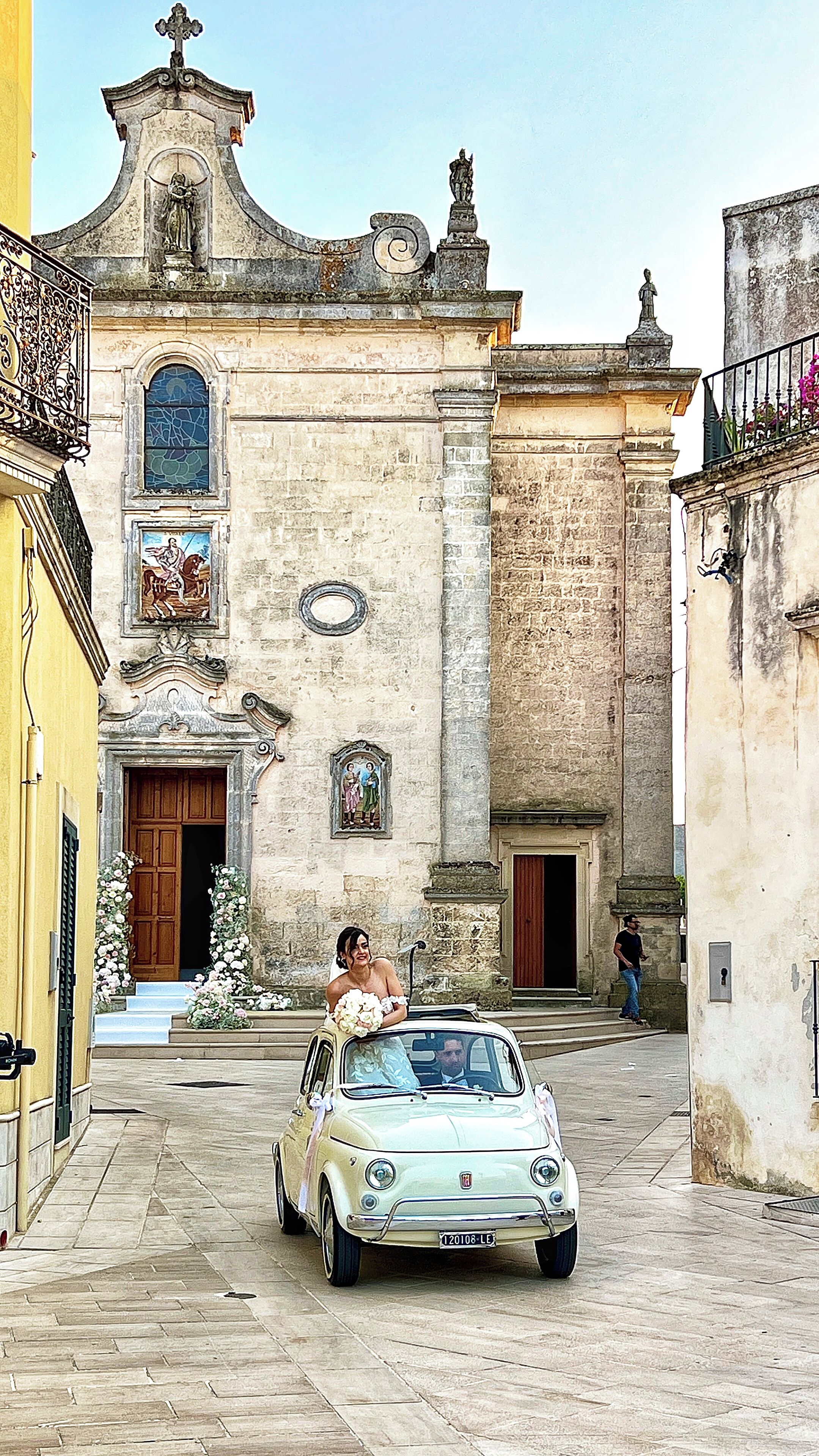 A vintage Fiat car parked in front of a historic building with a church facade, a balcony with flowers, and a statue on top.