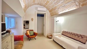 Une pièce avec un plafond en pierre, un canapé blanc, un fauteuil, un bureau avec un ordinateur et une fenêtre.
