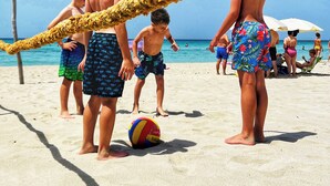 Une scène de plage où l’on voit des gens jouer au volleyball, une barrière faite de cordes et, au fond, des parasols.