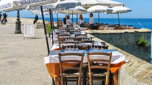 Tables de jardin avec des parasols blancs et des chaises en bois, offrant une vue sur la mer.