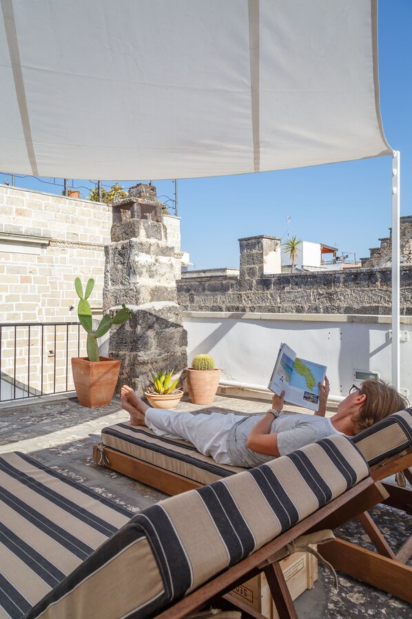 A person lying on a sunbed under a canopy, reading a book, with potted plants and a stone wall in the background.