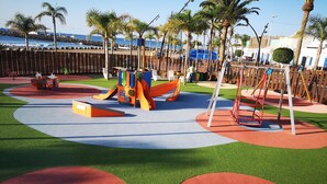 A playground with slides and swings, surrounded by palm trees and a view of the sea.