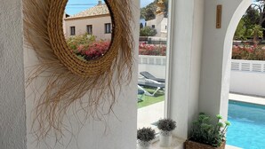Quarto com paredes brancas, um tapete de parede trançado, uma janela com vista para uma piscina e vegetação, e plantas em vasos.