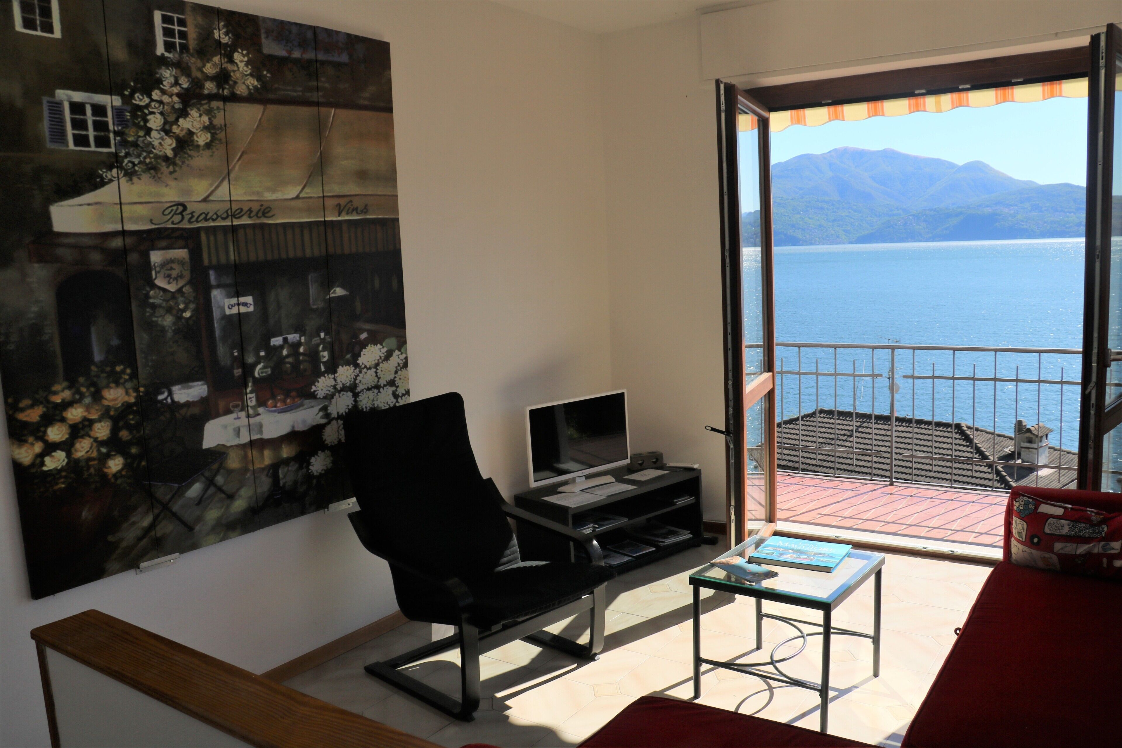 A room with a large painting of a restaurant, a black office chair, a small table with a laptop, and a view of a lake through a balcony.