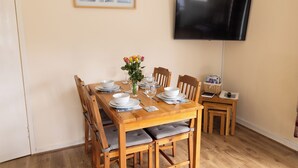 A dining area with a wooden table set for four, a vase with flowers, and a television mounted on the wall. - Central Lerwick holiday apartment (Lerwick)