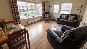 A living room with a wooden dining table set, a black leather sofa, and a window with curtains. - Central Lerwick holiday apartment (Lerwick)