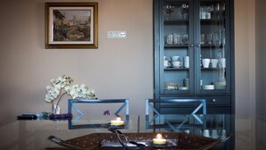 A dining area with a glass table, chairs, a candle, a framed painting, and a cabinet with glass doors.