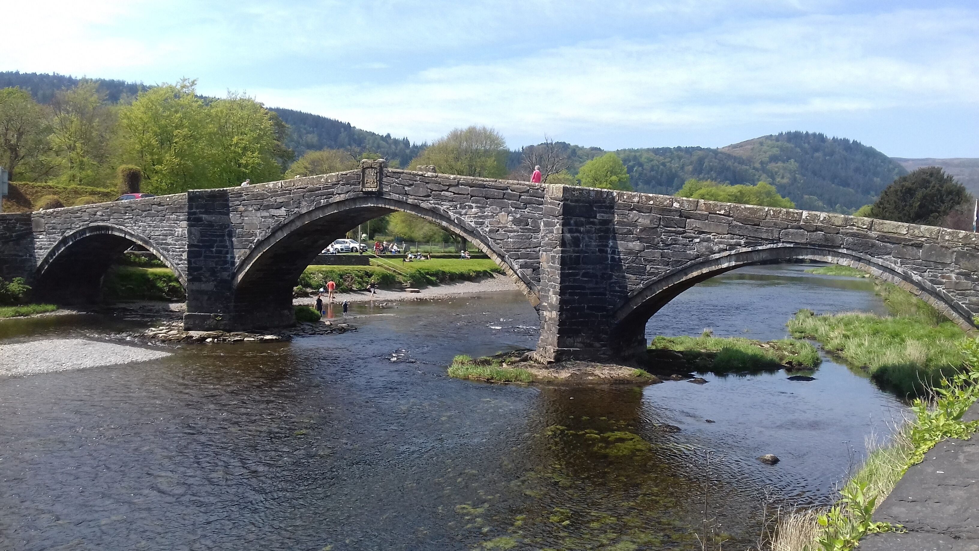 A stone bridge over a river with a grassy area and trees in the background.