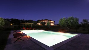 A swimming pool with a pergola and lounge chairs, surrounded by trees and a house with illuminated windows. - Villa Il Meluccio 12, Emma Villas (Castellina Marittima)