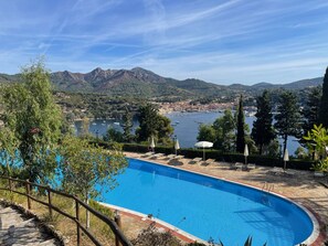 En swimmingpool med en terrass av sten, solstolar och parasoller. I bakgrunden syns en sjö med båtar och en stad på andra sidan vattnet.