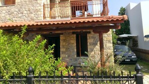 A stone house with a balcony, a car parked in the driveway, and a fence in the foreground.