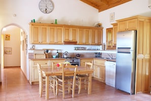 A kitchen with wooden cabinets, a stainless steel refrigerator, a dining table with chairs, and a clock on the wall. - Renovated traditional farmhouse overlooking the sea 400 olive trees.  (Nomos Ilias)