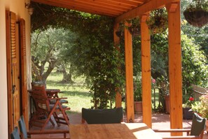 A covered patio with wooden furniture and a view of a lush garden. - Renovated traditional farmhouse overlooking the sea 400 olive trees.  (Nomos Ilias)