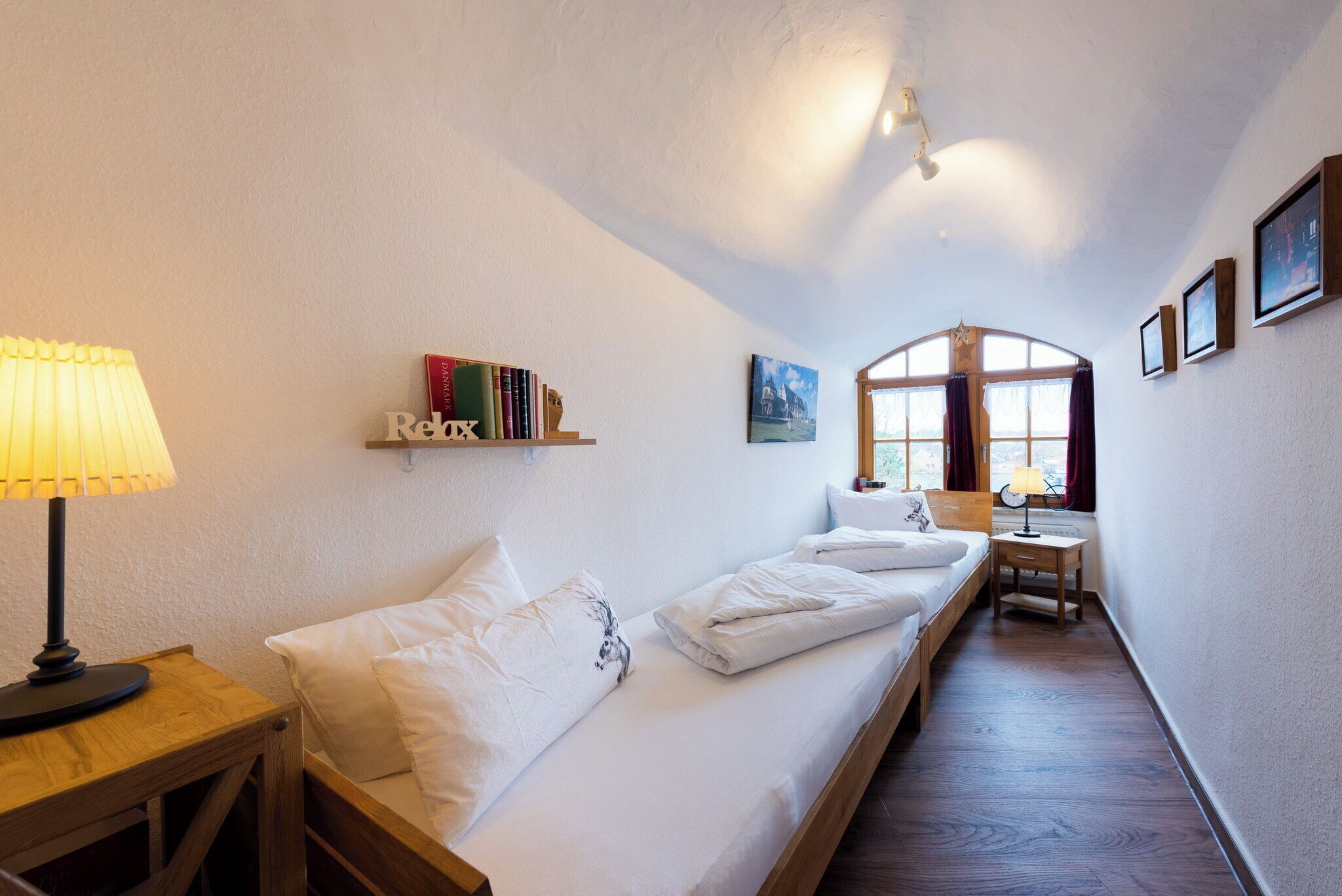 Una habitación pequeña y acogedora con una cama individual, una mesita de noche con lámpara, una estantería con libros y una ventana con cortinas.