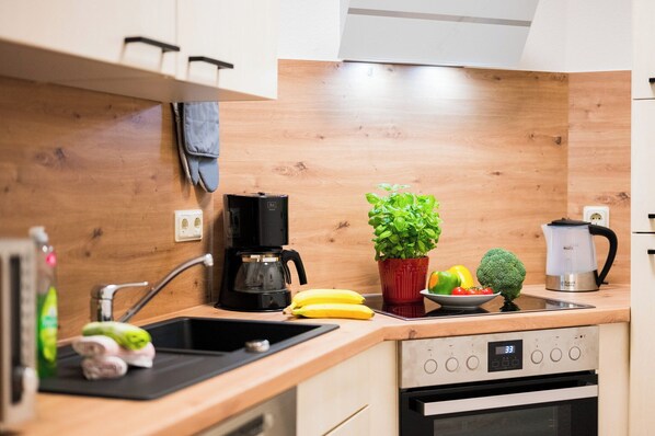 A modern kitchen with wooden cabinets, a black coffee maker, a red pot with a plant, a bowl of fruit, and a stainless steel kettle.
