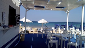 A beachside restaurant with outdoor seating, white umbrellas, and a view of the sea.