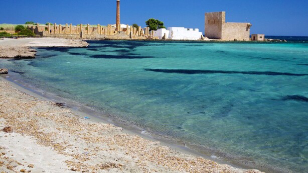 A coastal area with clear turquoise waters, sandy beach, and historical stone buildings.