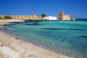 A coastal area with clear turquoise waters, sandy beach, and historical stone buildings.