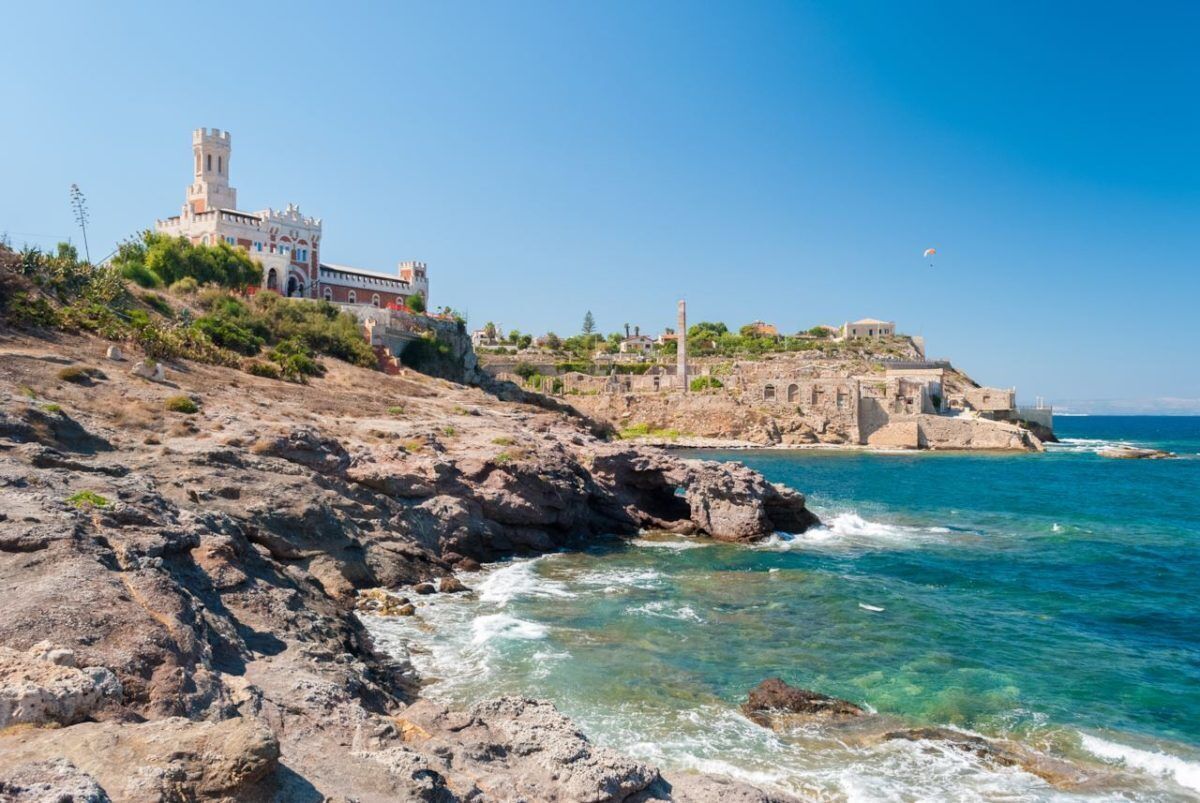 Un paysage côtier avec une structure ressemblant à un château, située sur une colline, un rivage rocheux et une mer bleue et limpide.