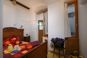 A bedroom with a wooden bed, a bedside table with a lamp, a window with curtains, and a small table with a vase of flowers. - Apartment La COLLEGIALE in the old town with free parking (Colmar)