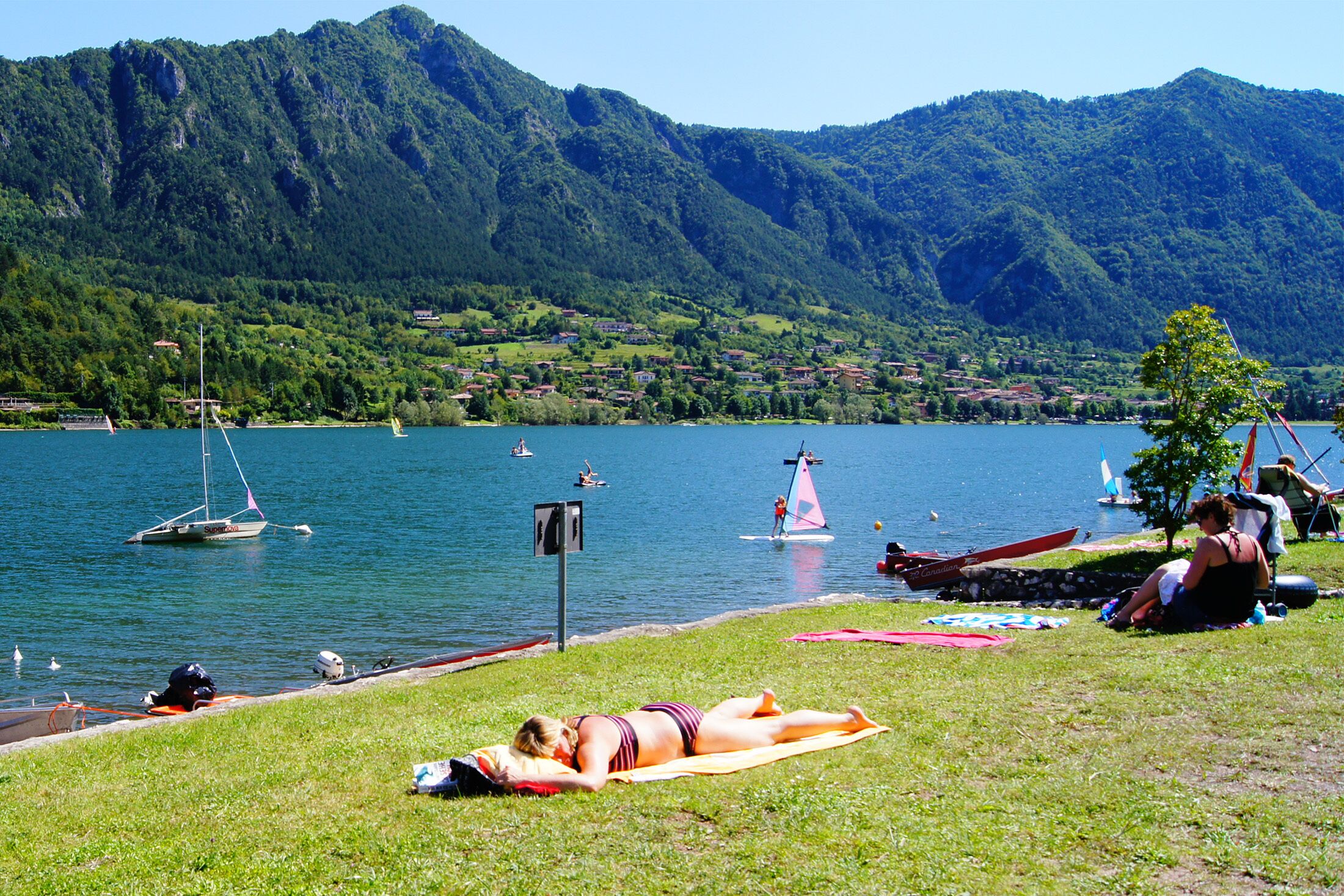 A lakeside view with boats, a woman sunbathing, and a mountainous backdrop.