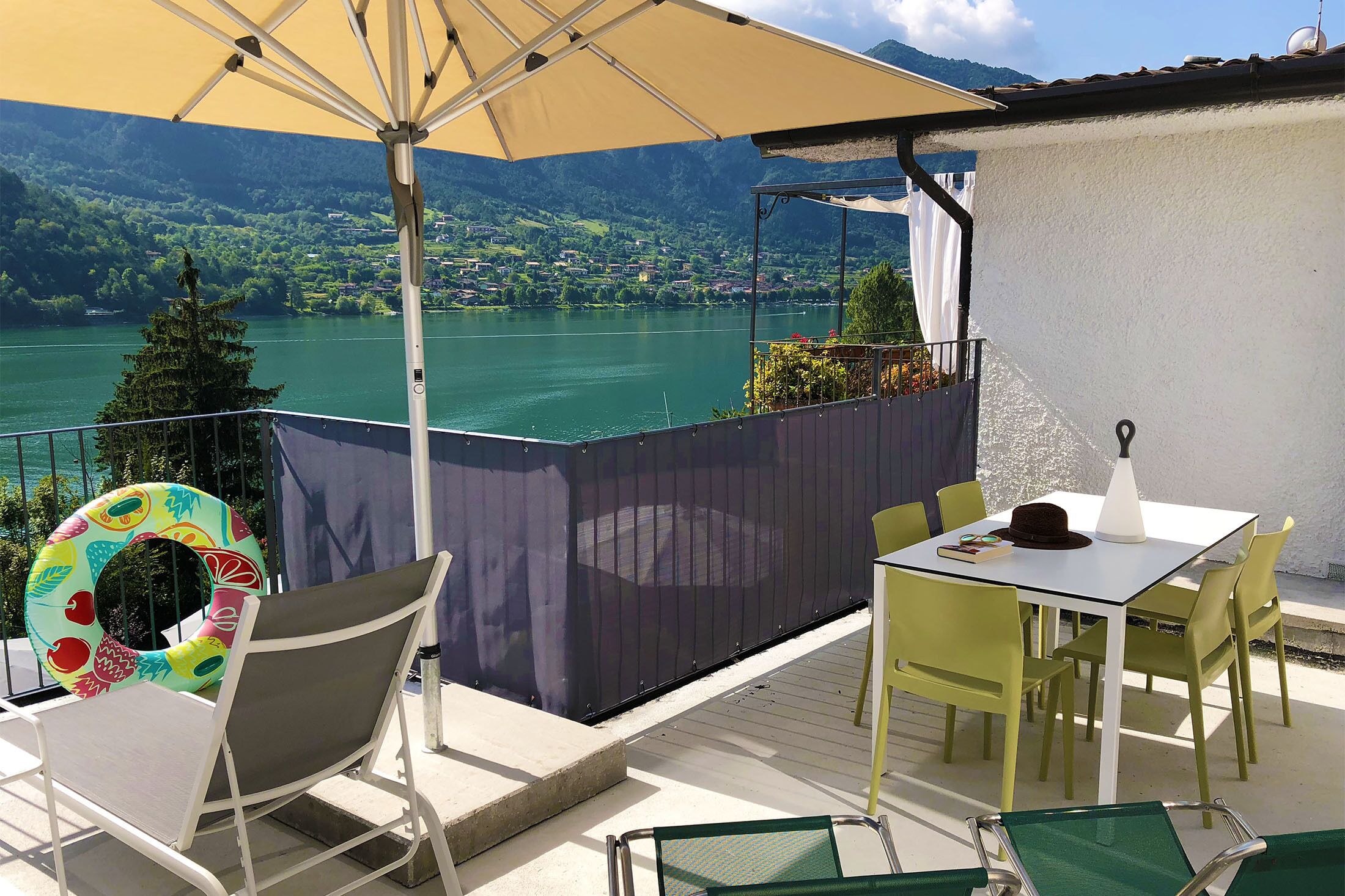 A terrace with a table and chairs, a poolside chair, and a view of a lake and mountains.
