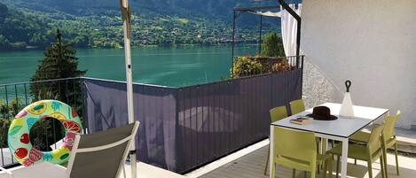 A terrace with a table and chairs, a poolside chair, and a view of a lake and mountains.