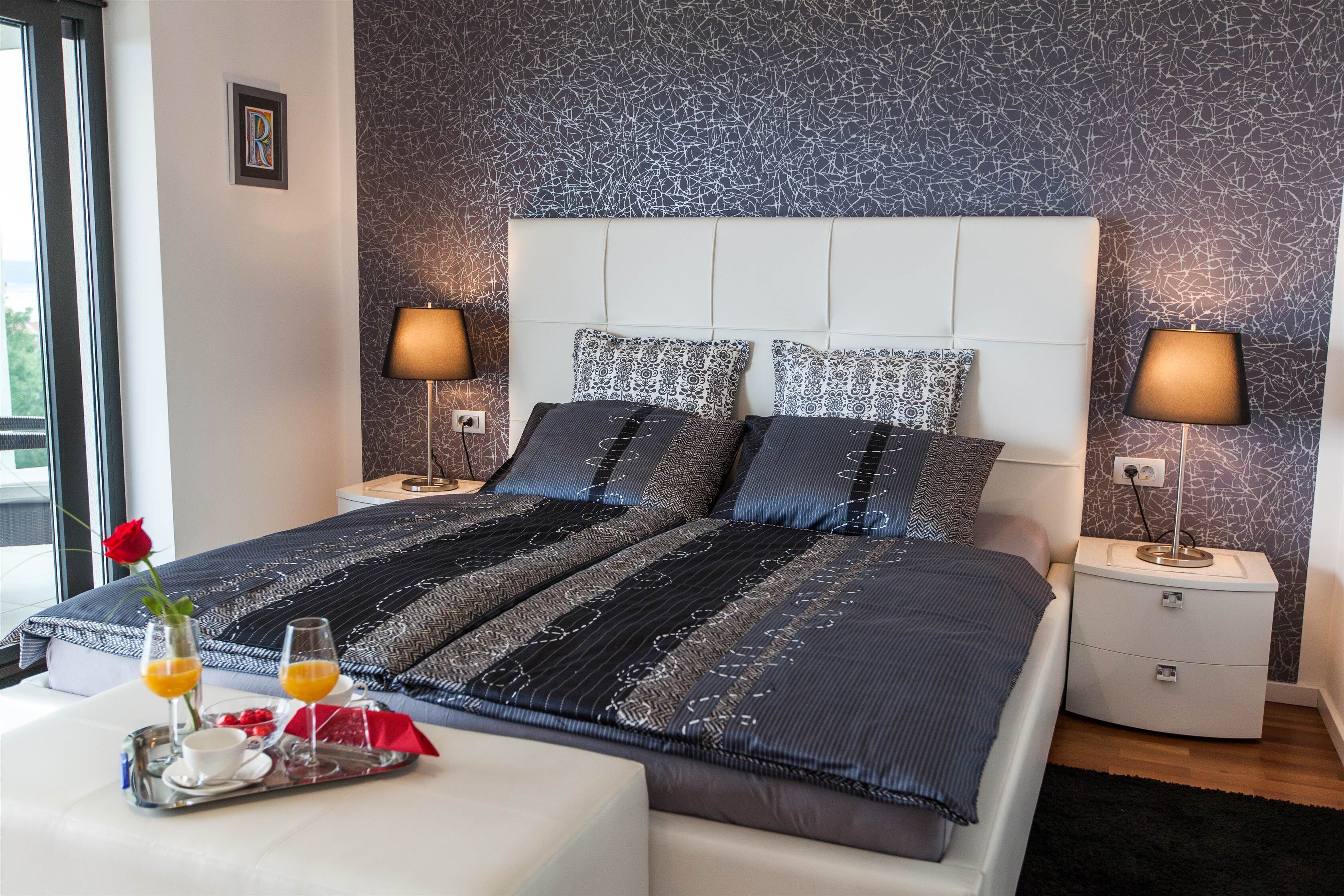 A neatly made double bed with a patterned bedspread, two bedside lamps, a tray with breakfast items, and a framed letter on the wall.