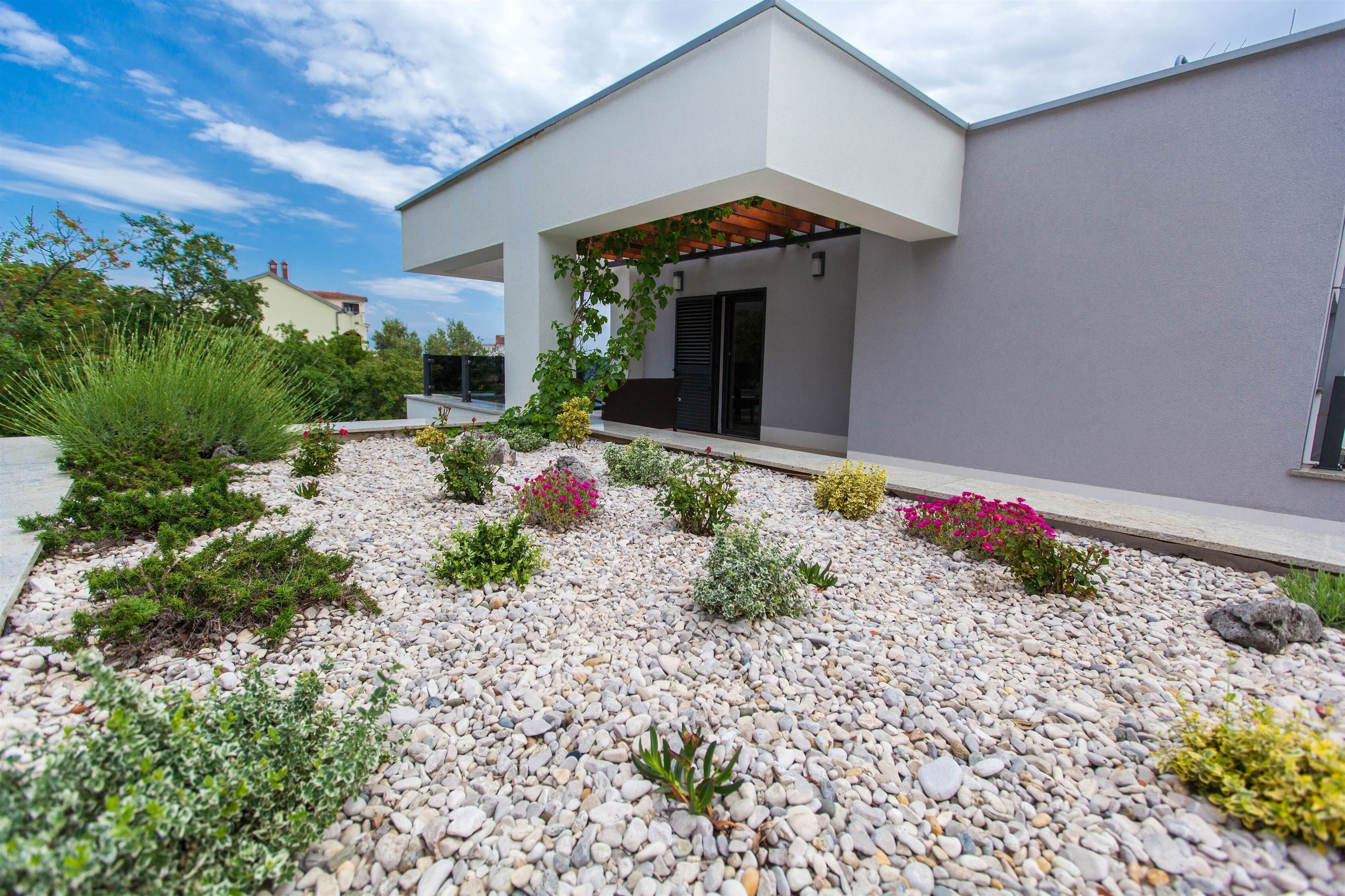 A modern house with a covered porch, surrounded by a gravel garden with various plants and flowers.