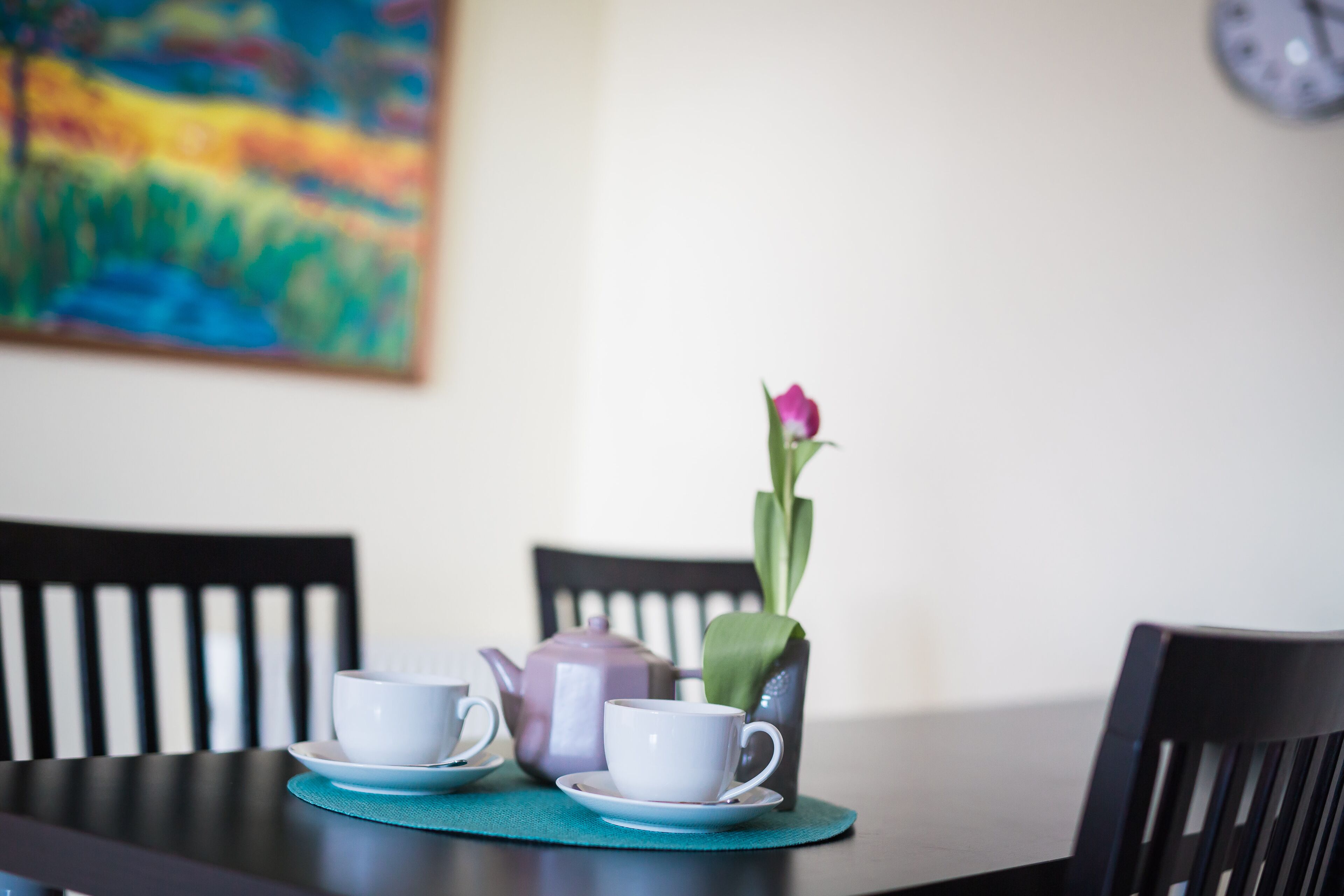 A dining table with two white cups and a teapot, a vase with a single flower, and a clock on the wall.