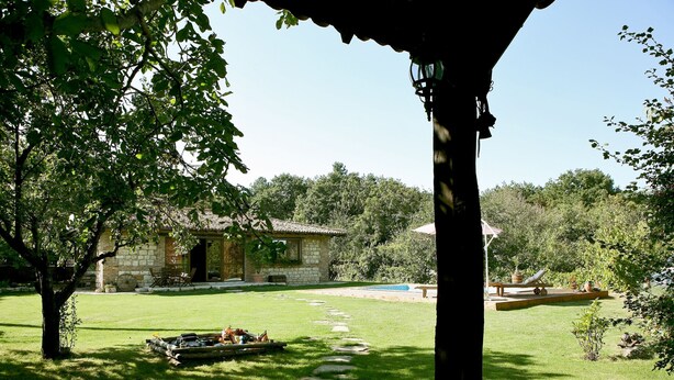 A stone house with a stone pathway leading to a pool area, surrounded by lush greenery and trees.