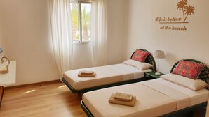 A room with two single beds, a window with sheer curtains, a wall decoration saying "Life is better at the beach", and a bedside table with a lamp.
