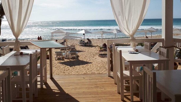 Un restaurant situé en bord de mer, avec des tables et des chaises blanches, une terrasse en bois et des parasols de plage.